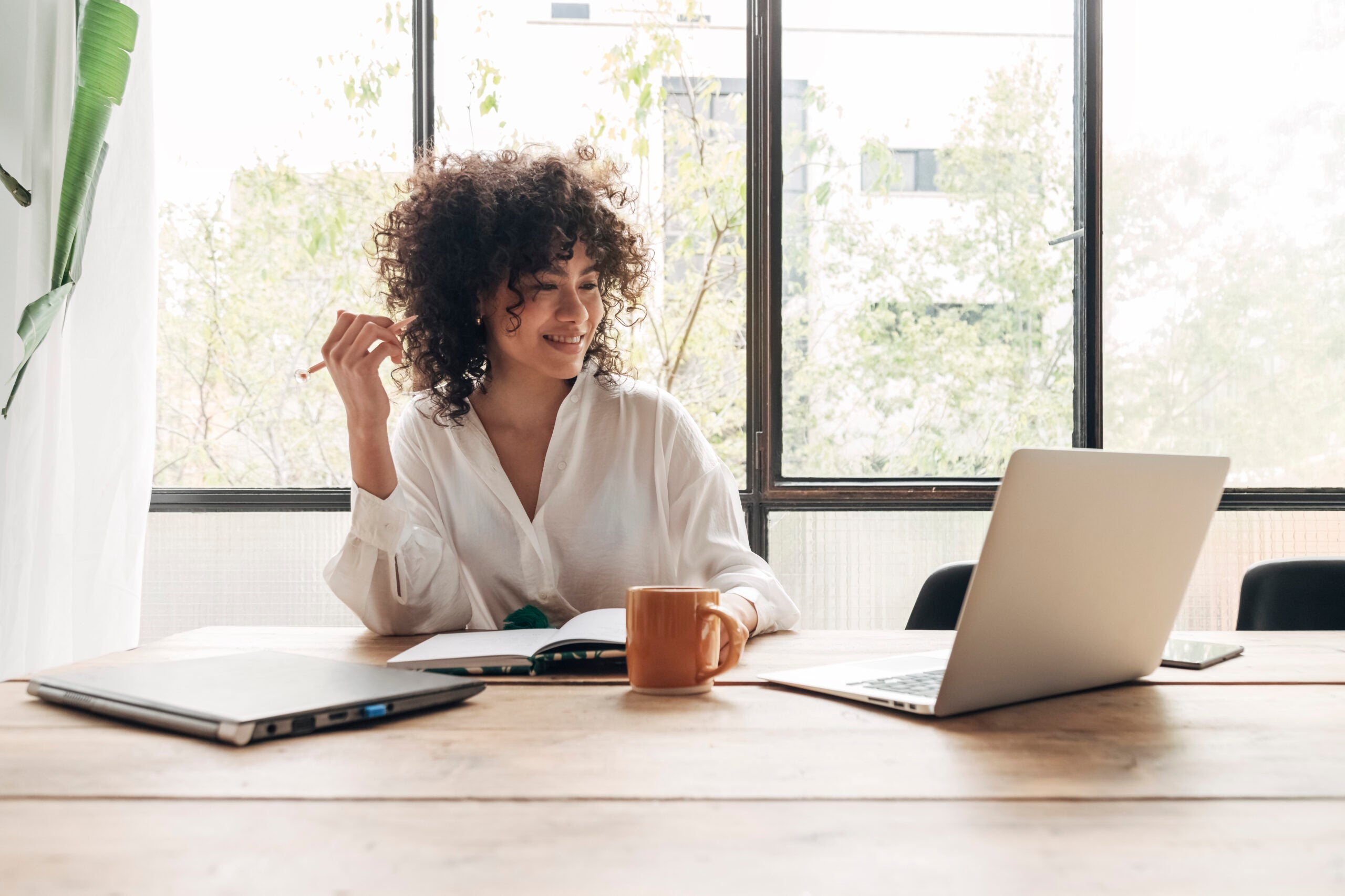 A woman is smiling and sitting at her desk with her laptop open while writing in her notebook.